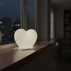 May include: A white, heart-shaped lamp on a wooden windowsill. The lamp is lit, emitting a soft glow. The background shows a cityscape at night, with blurred lights. The lamp is a decorative lighting fixture.