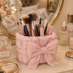 Può includere: Un portapennelli da trucco cilindrico, trapuntato e di colore rosa chiaro, caratterizzato da un grande fiocco decorativo sul davanti. Il contenitore è riempito con vari pennelli da trucco con manici metallici, scovolini per mascara e tubetti cosmetici rosa.