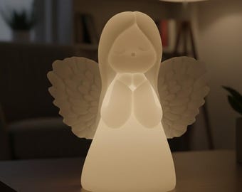Puede incluir: Una figura de ángel blanca y brillante con las manos juntas y alas detalladas. El ángel tiene los ojos cerrados y cabello largo. La lámpara está iluminada, emitiendo un brillo suave. El ángel es un objeto decorativo.