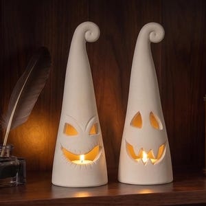 以下が含まれることがあります： オフホワイトの円錐形セラミック製キャンドルホルダー2点。彫り込まれたギザギザの表情が特徴です。各ピースは先端がカーブした尖った形状で、内部の光源が切り抜かれた目と大きく歯を見せた口を内側から照らしています。