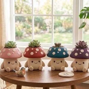 May include: Four mushroom-shaped succulent planters in pink, red, teal, and purple, each with white polka dots. The planters have smiling faces and hold small succulents. A small bird figurine and a coaster are on the wooden table.