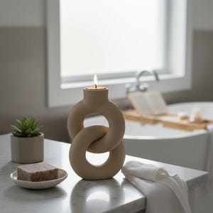 May include: A beige ceramic candle holder with a lit candle, featuring an interlocking ring design. A bar of soap on a small dish, a potted succulent, and a folded white towel are also visible on a marble surface. The scene is set in a bathroom.
