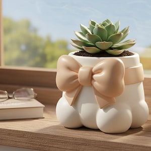 May include: A white ceramic plant pot with a bow detail, containing a green succulent. The pot has a distinctive shape with three rounded feet. The bow is a light tan colour. The plant pot is on a wooden surface.