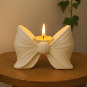 May include: A cream-coloured, bow-shaped candle holder with a lit tea light candle. The ceramic holder has detailed ribbing and a rounded centre. The flame of the candle is bright yellow. The candle holder is on a wooden surface.