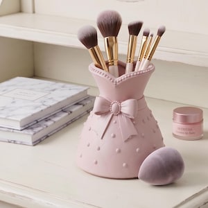 Può includere: Un portapennelli rosa a forma di vestito, ornato da un fiocco e dettagli a pois, contiene diversi pennelli per il trucco con manici dorati e setole morbide. Sono presenti anche una spugnetta per il trucco e un balsamo per le labbra.