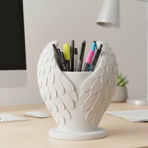 May include: White pen holder shaped like angel wings, filled with pens and markers of various colors. The desk organizer is made of a matte material and sits on a wooden desk. A computer monitor and lamp are in the background.