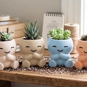 May include: Four small ceramic planters shaped like seated figures with smiling faces. Each planter holds a different succulent plant. The planters are colored in soft shades of light pink, beige, light blue, and peach, featuring arms crossed.