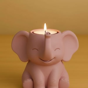 Könnte beinhalten: Ein rosa elefantenförmiger Kerzenhalter mit einer brennenden Kerze im Inneren. Der Kerzenhalter hat große Ohren, einen Rüssel und ein lächelndes Gesicht. Der Kerzenhalter besteht aus einem matten Material und steht auf einer gelben Oberfläche.