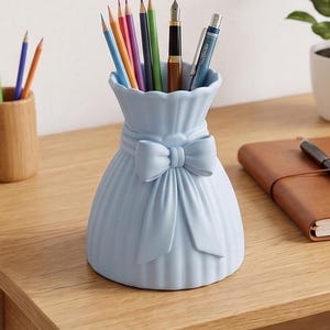 Può includere: Un portapenne in ceramica azzurra a forma di sacchetto con fiocco. Contiene matite colorate, una penna e una matita meccanica. È appoggiato su una scrivania di legno, accanto a un quaderno e una pianta.