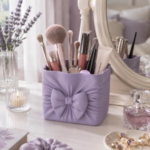 Op de afbeelding: Een vierkante lavendelkleurige make-up kwastenhouder met een gebeeldhouwde strik aan de voorzijde en een geplooide textuur. De houder is gevuld met diverse make-up kwasten, lipgloss tubes en een kleine tube crème met zichtbare tekst.