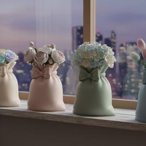 Puede incluir: Cuatro jarrones decorativos con forma de bolsa, cada uno con un lazo y llenos de flores. Los jarrones son de colores pastel: crema, rosa, verde y azul. Las flores incluyen rosas y tulipanes, creando una exhibición suave y elegante.