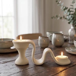 Puede incluir: Un portavelas de cerámica blanca mate con una forma continua, fluida y ondulada que conecta dos plataformas circulares. Cada plataforma sostiene una pequeña vela de té encendida con una llama visible, creando una pieza de iluminación.