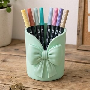 Può includere: Un portapenne cilindrico color verde menta con un fiocco sagomato in rilievo sulla parte anteriore. Il supporto contiene otto pennarelli a doppia punta in vari colori pastello, tra cui giallo, corallo, azzurro, ottanio, pervinca, rosa, viola e beige.