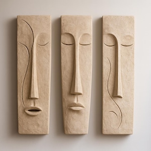 Może przedstawiać: Trzy prostokątne elementy sztuki ściennej z wyrzeźbionymi twarzami. Każdy element ma długi nos, zamknięte oczy i usta. Sztuka jest w neutralnym beżowym kolorze, z subtelną fakturą i jest zamontowana na białej ścianie.