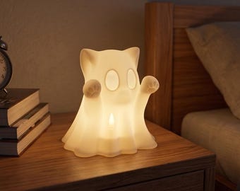 May include: A white, translucent, ghost-shaped lamp with cat-like ears and raised paws featuring visible paw pads. The lamp emits a soft, warm glow from within, illuminating its hollow interior and smooth, rounded form on a wooden surface.