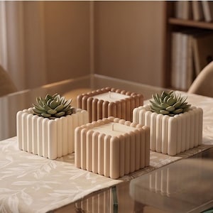 May include: Four square decorative planters with a ribbed design. Two planters hold small succulents, and two hold candles. The planters are in shades of beige and brown, arranged on a glass table with a floral runner.