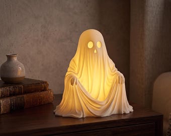 May include: A white, ghost-shaped lamp with a smooth, matte finish and a warm, glowing interior. The lamp features two circular eye cutouts and a small triangular mouth cutout, emitting a soft, diffused light from within its hollow structure.