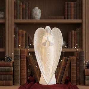 May include: A white angel ornament with open wings and hands clasped in prayer, lit from within. It stands on a red cloth, in front of a bookshelf filled with old books. The angel is a decorative item, suitable for a home setting.