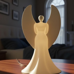 Pode incluir: Uma lâmpada branca em forma de anjo, com um interior brilhante e grandes asas curvas. A lâmpada está numa mesa de madeira, com um cabo que sai da sua base. O fundo inclui uma cama e quadros emoldurados, sugerindo um quarto.