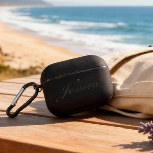 Könnte beinhalten: Schwarzes AirPods-Etui mit dem eingravierten Namen "Jessica" auf der Vorderseite. Das Etui hat einen silbernen Karabinerhaken. Das Etui liegt auf einem Holztisch mit Strand und Meer im Hintergrund.