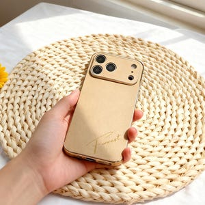 Op de afbeelding: Een goudkleurige telefoonhoes met een camera-uitsparing en het woord "Forrest" in cursief schrift. De hoes wordt in een hand gehouden en rust op een geweven, ronde placemat. Op de achtergrond is een gele bloem te zien.