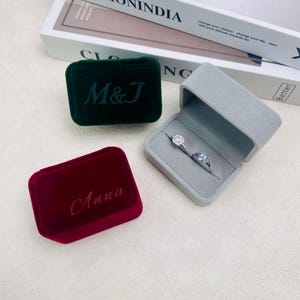 May include: Three velvet ring boxes in green, burgundy, and gray. The green box has "M&J" embroidered on it, the burgundy box has "Anna" embroidered on it, and the gray box is open, displaying two rings.