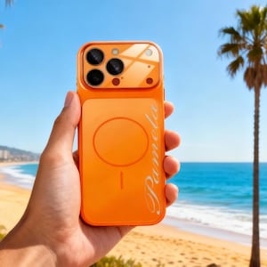 Puede incluir: Funda de teléfono naranja brillante con un módulo de cámara y el nombre "Pamela" en cursiva. La funda tiene un diseño circular en la parte trasera. El fondo muestra una playa, una palmera y cielo azul.