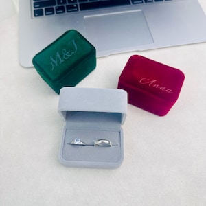 May include: A light grey ring box containing two rings, alongside a green velvet box embroidered with "M & J" and a red velvet box embroidered with "Anna". The rings include a silver band and a ring with a square gemstone.