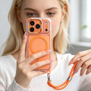 May include: A clear and orange phone case with a built-in circular magnetic charging ring on the back. The case features a metallic orange camera lens frame and includes a detachable orange cord lanyard attached to the bottom corner.