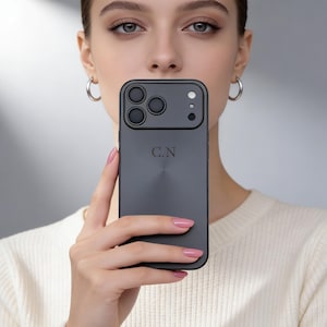 Puede incluir: Una funda de teléfono gris oscuro con acabado mate, con tres lentes de cámara y una sola lente. La funda tiene las iniciales "C.N" grabadas en la parte posterior. La funda del teléfono está diseñada para proteger el teléfono.
