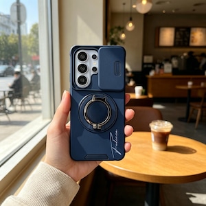 May include: A navy blue phone case with a camera cover and a gold ring holder. The case has a smooth finish and the name "Felicia" is written in a cursive font on the lower right corner.