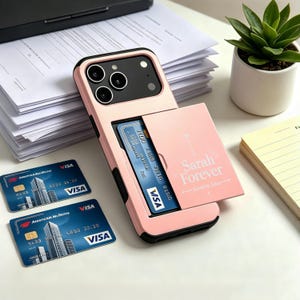 Puede incluir: Una funda de smartphone de color rosa claro con ranura para tarjetas integrada. La funda cuenta con una tapa deslizante impresa con el texto Sarah Forever Loved by John. Se ven dos tarjetas de crédito Visa azules, una dentro y otra al lado.