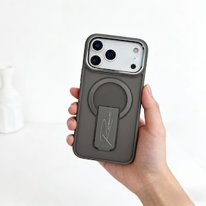 May include: A dark grey phone case with a silver ring holder. The case has a circular cutout for the camera and a silver frame. The ring holder has the word "Rorenzo" printed on it. The phone case is held in a person's hand.