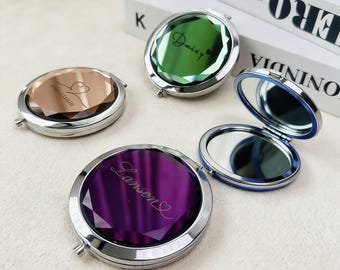 May include: Four compact mirrors with a faceted design. One mirror is open, revealing a reflective surface with a blue border. The other mirrors have colored surfaces in shades of brown, green, and purple, each with a name engraved.