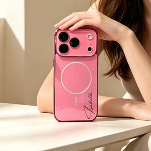 Puede incluir: Una funda de smartphone de color rosa brillante con un anillo de carga magnético circular y una línea de alineación vertical. La funda tiene un recorte para tres lentes de cámara y un flash. El nombre Isabella está impreso en cursiva negra abajo.