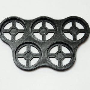 May include: A black plastic tray with six circular cutouts, each with a cross-shaped design. The tray is designed to hold small round objects.