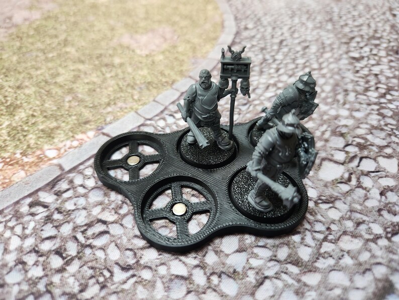 May include: A black plastic base with four circular slots for miniature figures. Each slot has a magnetic center and a raised rim. Three miniature figures are displayed on the base, two in armor and one with a banner.