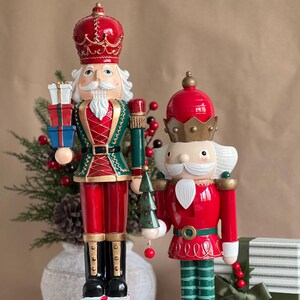 Op de afbeelding: Twee decoratieve kerstnotenkrakers. De grotere notenkraker is rood, groen en goud, met cadeaus en een staf. De kleinere notenkraker is rood en groen, met een kerstboom. Beide hebben wit haar en kronen.