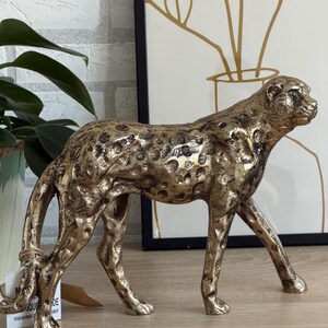 May include: A gold-coloured cheetah figurine with black spots. The sculpture is detailed, with visible muscles and a textured surface. The cheetah is in a walking pose, with its head turned to the side. The figurine is a decorative item.