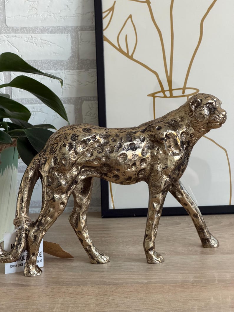 May include: A gold-coloured cheetah figurine with black spots. The sculpture is detailed, with visible muscles and a textured surface. The cheetah is in a walking pose, with its head turned to the side. The figurine is a decorative item.