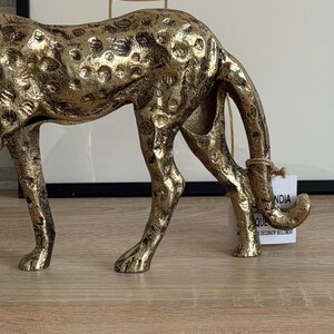 May include: A gold-coloured, decorative cheetah figurine with a textured surface. The cheetah is in a crouching position, with its head lowered. The figurine has black spots and is made of metal.