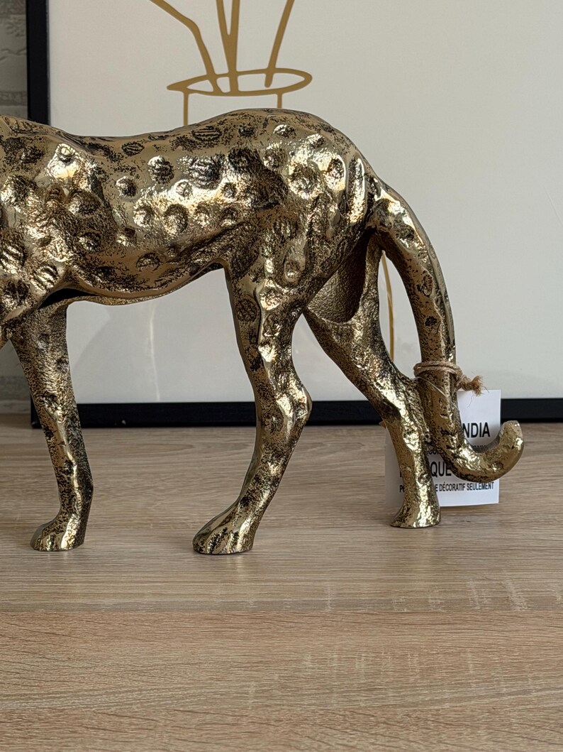 May include: A gold-coloured, decorative cheetah figurine with a textured surface. The cheetah is in a crouching position, with its head lowered. The figurine has black spots and is made of metal.