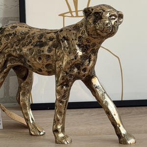 May include: A gold-coloured cheetah figurine with a textured surface and black spots. The cheetah is in a walking pose, with its head turned slightly to the right. The figurine is made of metal and is a decorative item.