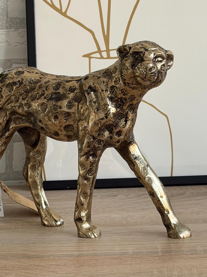 May include: A gold-coloured cheetah figurine with a textured surface and black spots. The cheetah is in a walking pose, with its head turned slightly to the right. The figurine is made of metal and is a decorative item.