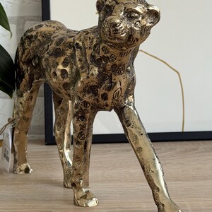 May include: A gold-coloured leopard statue with a textured surface and dark brown spots. The sculpture is in a walking pose, with its head turned slightly to the right. The statue is a decorative home accent.