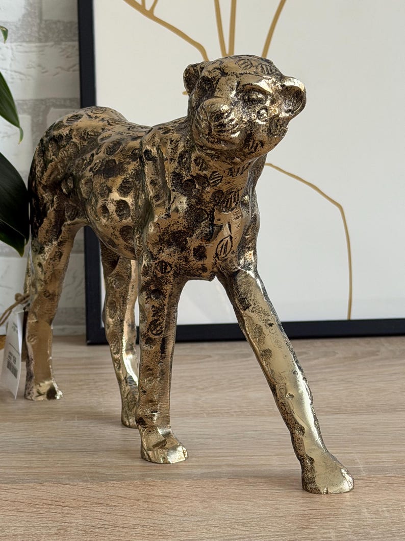 May include: A gold-coloured leopard statue with a textured surface and dark brown spots. The sculpture is in a walking pose, with its head turned slightly to the right. The statue is a decorative home accent.