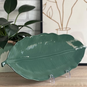 Puede incluir: Una fuente verde de cerámica con forma de hoja. La fuente tiene vetas detalladas y un acabado brillante. Se exhibe sobre una superficie de madera con soportes de plástico transparente. Una planta en maceta y una impresión botánica enmarcada están en el fondo.