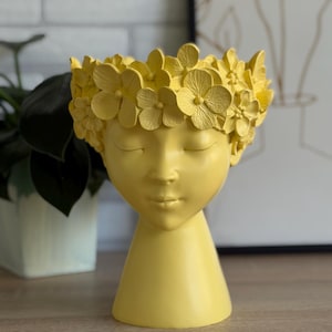 Könnte beinhalten: Ein gelber dekorativer Pflanzentopf in Form eines ruhigen Gesichts, geschmückt mit einem Blumenkranz. Der Topf hat geschlossene Augen und eine glatte Oberfläche. Der Sockel hat eine konische Form. Der Hintergrund umfasst eine Pflanze und ein gerahmtes Kunstwerk.