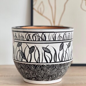 Könnte beinhalten: Ein weißer Keramik-Pflanztopf mit einem schwarzen Blatt- und Stiel-Design. Der Topf hat eine weite Öffnung und eine abgerundete Form. Der Rand und der Boden weisen detaillierte schwarze Muster auf. Der Innenraum ist terrakottafarben.