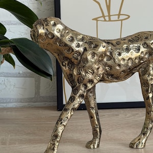 May include: A gold cheetah sculpture with black spots. The decorative figurine is made of metal and has a textured surface. The cheetah is posed in a walking position, with its head turned to the side. The sculpture is a home decor item.